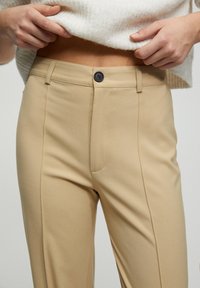 Pantalons taillés beiges avec une texture lisse, dotés d'un bouton noir et de détails de couture sur les côtés, présentés avec un pull léger.
