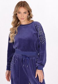 Lila velourtröja med långa ärmar, prydd med silvernitar på axlarna, ribbade ärmslut och en matchande midja.