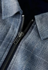 helehelesinine teksariie, millel on lähedalt vaadatav metallist tõmblukk, millel on hõbedane tõmbetihvt, koos kraega kaelusega.