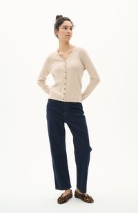 Cardigan beige a costine con scollatura a V e chiusura con bottoni, abbinato a jeans a gamba ampia blu scuro e pantofole con stampa leopardata.