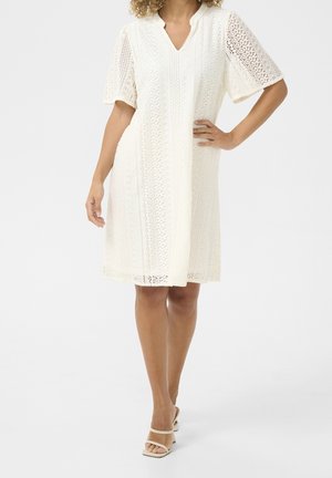 Femme portant une robe blanche en dentelle longueur genou avec manches courtes et col en V, associée à des sandales ouvertes crème à talons.