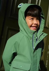 Grön regnrock med grodtema på huvan, utrustad med runda ögon, dragkedja, tryckknappar och framficka. Slät yta, vattentätt material.