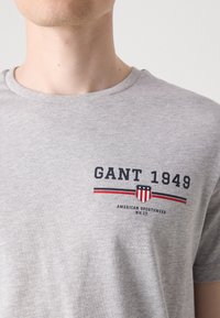 Szürke pamut póló kerek nyakkivágással, "GANT 1949" nyomott logóval, piros és sötétkék kiegészítésekkel. Simább textúra, laza fazon.