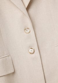 Beige wollen blazer met twee grote doorschijnende knopen en een ingezette revers, waarbij een deel van een flapzak aan de linkerkant zichtbaar is.