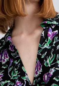 Černá květinová blůza s zelenými a fialovými vzory. Má límec, krátké rukávy a hluboký výstřih do V. Měkká textura.
