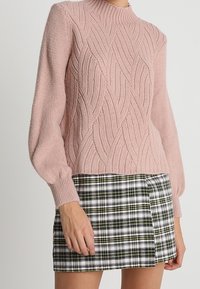 Różowy sweter na drutach z fakturą falistego wzoru, wysokim kołnierzem i bufiastymi rękawami, zestawiony z mini spódniczką w kratę w odcieniach zieleni i czerni.