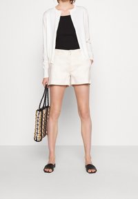 Kremowy kardigan, czarny top na ramiączkach, jasnobeżowe szorty oraz czarne sandały wsuwane; trzyma plecioną torbę na ramię w kolorach czarnym, białym i brązowym.