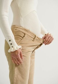 Beige Cordhosen mit lockerer Passform und seitlichem elastischen Einsatz. Weißes langärmeliges Shirt mit gerippter Struktur, Rüschen an den Manschetten und Knöpfen.