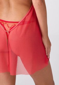 Czerwona siateczkowa koszulka nocna z koronkowymi akcentami, z dekoltem w serek i odkrytymi plecami. Miękka w dotyku i lekka tkanina podkreśla kobiece kształty.