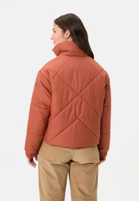Roodbruine pufferjack met een hoge kraag, een diagonaal quiltingpatroon en elastische manchetten, gecombineerd met beige corduroy broek.