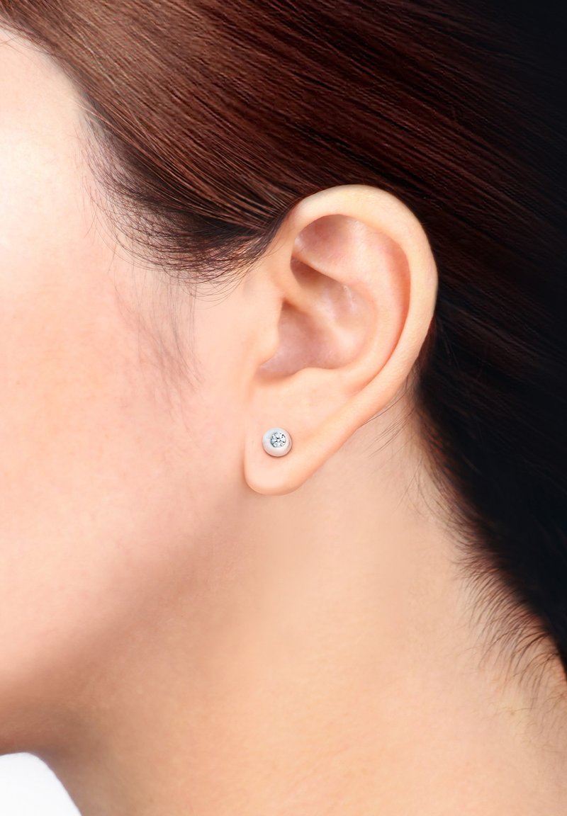 Silver studs med ett cirkulärt mönster och en klar ädelsten i mitten, placerade mot en modellens öra med släta hår.