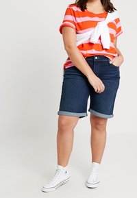 Camisa de manga corta a rayas naranja y rosa con un detalle atado en blanco, combinada con pantalones cortos de mezclilla oscura y zapatillas altas blancas.