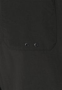 Vêtement noir avec un détail de poche plate et deux œillets en métal. Le matériau semble lisse avec une finition mate.