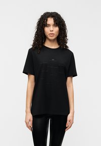 Μαύρο βαμβακερό T-shirt με στρογγυλή λαιμόκοψη, κοντά μανίκια και ανάγλυφο κείμενο σε διάφορα μεγέθη στο μπροστινό μέρος. Συνδυάζεται με γυαλιστερό μαύρο κολάν.