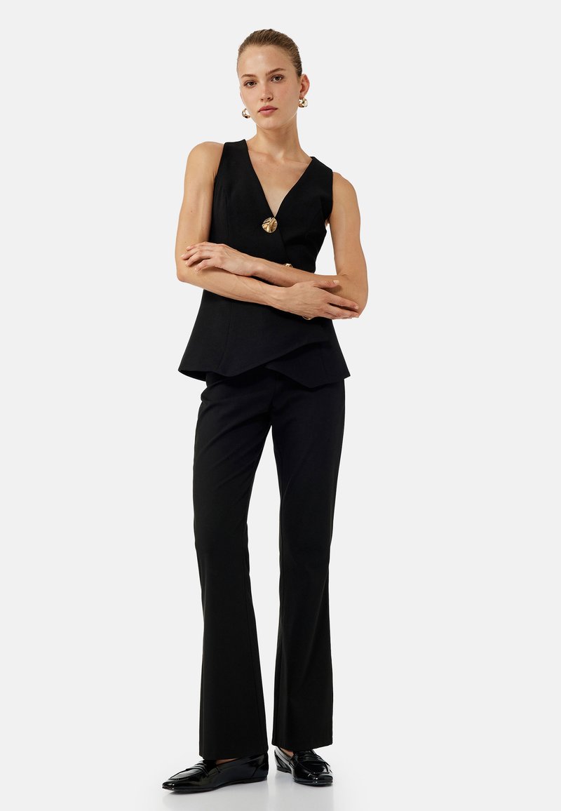Top peplum nero con scollo a V e orlo asimmetrico, abbinato a pantaloni neri svasati e mocassini neri lucidi. Dettaglio in metallo dorato sul top.