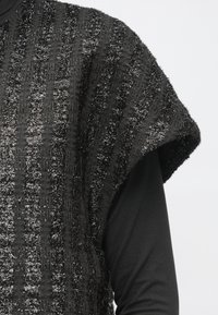 Crna i siva teksturirana bezrukava odjeća preko crne majice s dugim rukavima na osobi, prikazana od ramena do sredine trupa.