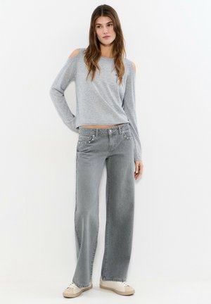 Mujer con suéter gris de hombros descubiertos, jeans grises de tiro alto y pierna ancha, y zapatillas blancas, posando frente a un fondo blanco.