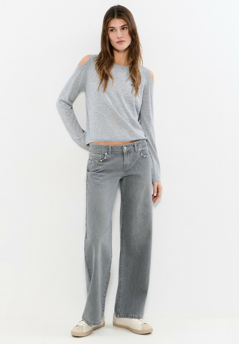 Mujer con suéter gris de hombros descubiertos, jeans grises de tiro alto y pierna ancha, y zapatillas blancas, posando frente a un fondo blanco.