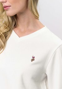 Vit bomullströja med V-ringning, med en broderad logotyp på vänster bröst. Mjuk textur och avslappnad passform.