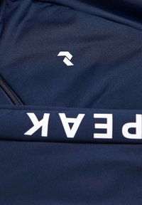 Mørkeblå stof med et hvidt geometrisk logo og fed, stiliseret tekst, der står "peak." Glat overflade med lynlåsdetalje.