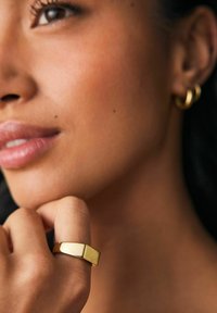 Goldener Metallring mit rechteckiger Form, glatter Oberfläche und matter Oberfläche, am Finger getragen, kombiniert mit einem runden Creolenohrring.