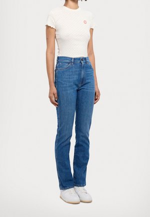 Femme portant un haut blanc à manches courtes avec un petit logo rouge, un jean bleu taille haute et des baskets blanches, debout devant un fond uni.