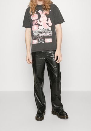 Camiseta gráfica negra con diseño frontal que presenta ilustraciones en rosa; combinada con pantalones de cuero negro brillantes y botas negras.