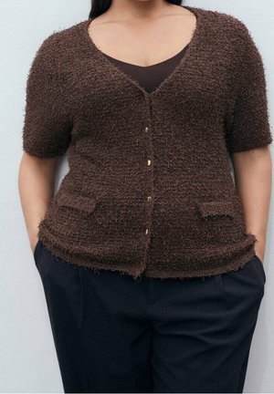 Femme portant un cardigan marron à boutons à manches courtes en tissu duveteux, par-dessus un haut sombre, les mains dans les poches d'un pantalon noir, sur un fond clair.