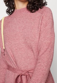 Maglione rosa lavorato a maglia con scollatura a costine. Presenta un dettaglio con cintura legata e una texture morbida. Maniche lunghe con vestibilità rilassata.