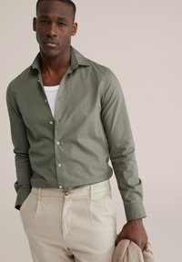 Dolga srajca svetlo olivno zelene barve z ovratnikom na gumbe, kombinirana s kremasto belimi hlačami. Srajca ima gladko teksturo in kontrastne gumbe.