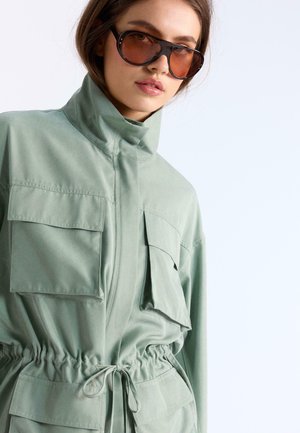 Frau trägt große, braun getönte Sonnenbrillen und eine hellgrüne Jacke mit mehreren Klappentaschen und einem Taillenbindegürtel.