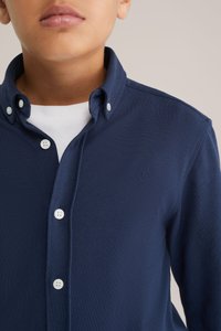 Tumši zila krekls ar garām piedurknēm, ar pogām sakņotu apkakli un mazu logotipu uz krūšu daļas. Izgatavots no teksturēta auduma, ar baltām pogām.