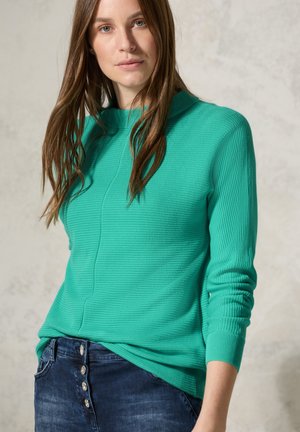 Green ribbed sweater with a high neck, featuring long sleeves and a textured finish, paired with dark denim jeans featuring button details.