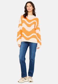 Oranje en roze golfpatroon gebreide trui met een ronde hals, ontspannen pasvorm en lange mouwen, gecombineerd met blauwe jeans en sneakers.