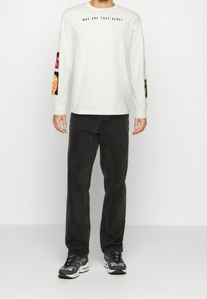 Hombre con camisa blanca de manga larga con texto "¿POR QUÉ ESTÁN AQUÍ?" y parches coloridos en las mangas, pantalones vaqueros negros y zapatillas negro-gris.