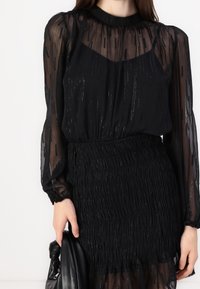 Svart genomskinlig blus med texturerade volanger, långa ärmar och en samlad midja. Har glittrande detaljer och en rund halsringning.