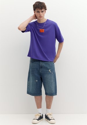 Jeune homme portant un T-shirt violet avec un patch orange, un short en jean, des chaussettes blanches et des baskets noires, debout contre un mur blanc.