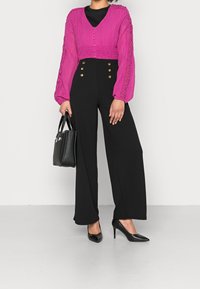 Roze gestructureerde cropped blouse met knoopsluiting, gecombineerd met zwarte wijde broek met gouden knopen. Zwarte handtas en hoge hakken.