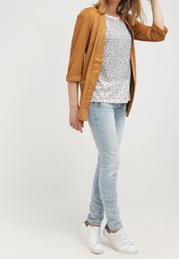 Senapsgul lättviktsjacka, vit topp med små svarta prickar, ljusblå slim fit-jeans och vita sneakers med mönstrade accenter.