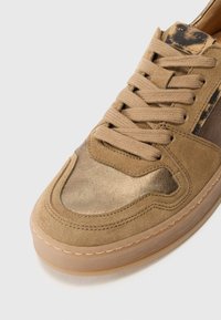Tan suede sneaker featuring a metallic gold toe, lace-up design, and a translucent rubber sole, with stitching details and minimal accents.