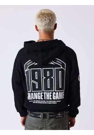 Personne aux cheveux blonds portant un sweat à capuche noir avec un grand texte blanc "1980 Change The Game" et un texte plus petit en dessous dans le dos.