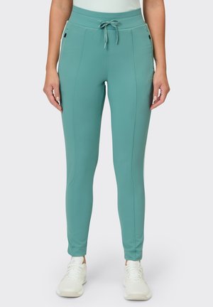 Frau trägt eng anliegende Jogginghosen in Petrol mit Kordelzug am Bund und Seitentaschen, kombiniert mit weißen Sneakers.
