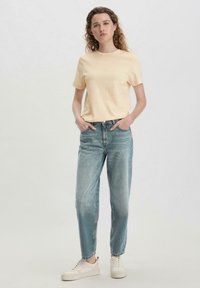 T-shirt di cotone giallo chiaro con scollo rotondo, abbinata a jeans a vita alta azzurri e sneakers bianche, in posa eretta.