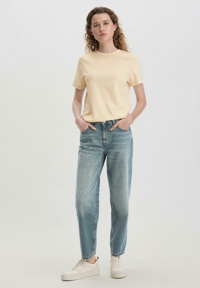 T-shirt di cotone giallo chiaro con scollo rotondo, abbinata a jeans a vita alta azzurri e sneakers bianche, in posa eretta.