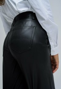 Persona vistiendo pantalones de piel sintética negros con costuras en los bolsillos traseros y una camisa blanca de manga larga, con la mano relajada al lado.