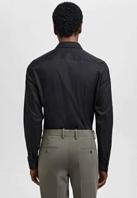 Cămășă neagră cu mâneci lungi, cu o textură netedă, cu mâneci cu butoni, asociată cu pantaloni eleganți gri deschis, cu buzunare la spate și curea.