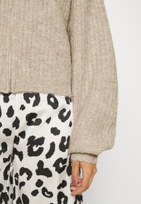 Beige gebreide cardigan met een rits aan de voorkant, met ribbeltextuur en oversized mouwen, gecombineerd met broek in zwart-wit luipaardprint.