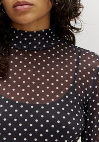 Schwarze, durchsichtige Bluse mit kleinen weißen Punkten, ausgestattet mit einem hohen Kragen und einer glatten Textur. Das Design umfasst lange Ärmel und einen runden Ausschnitt.