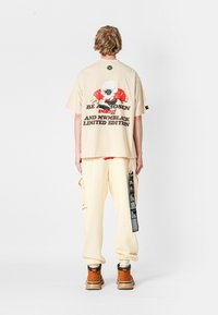 Camiseta oversized beige con un diseño gráfico que presenta un personaje de dibujos animados, texto negro y acentos rojos. Combinada con pantalones cargo crema y botas marrones.