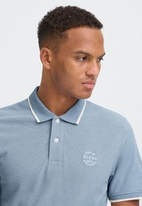 Bărbat cu păr scurt și creț, purtând un tricou polo bleu cu guler și margini albe, privind spre dreapta pe un fundal simplu.